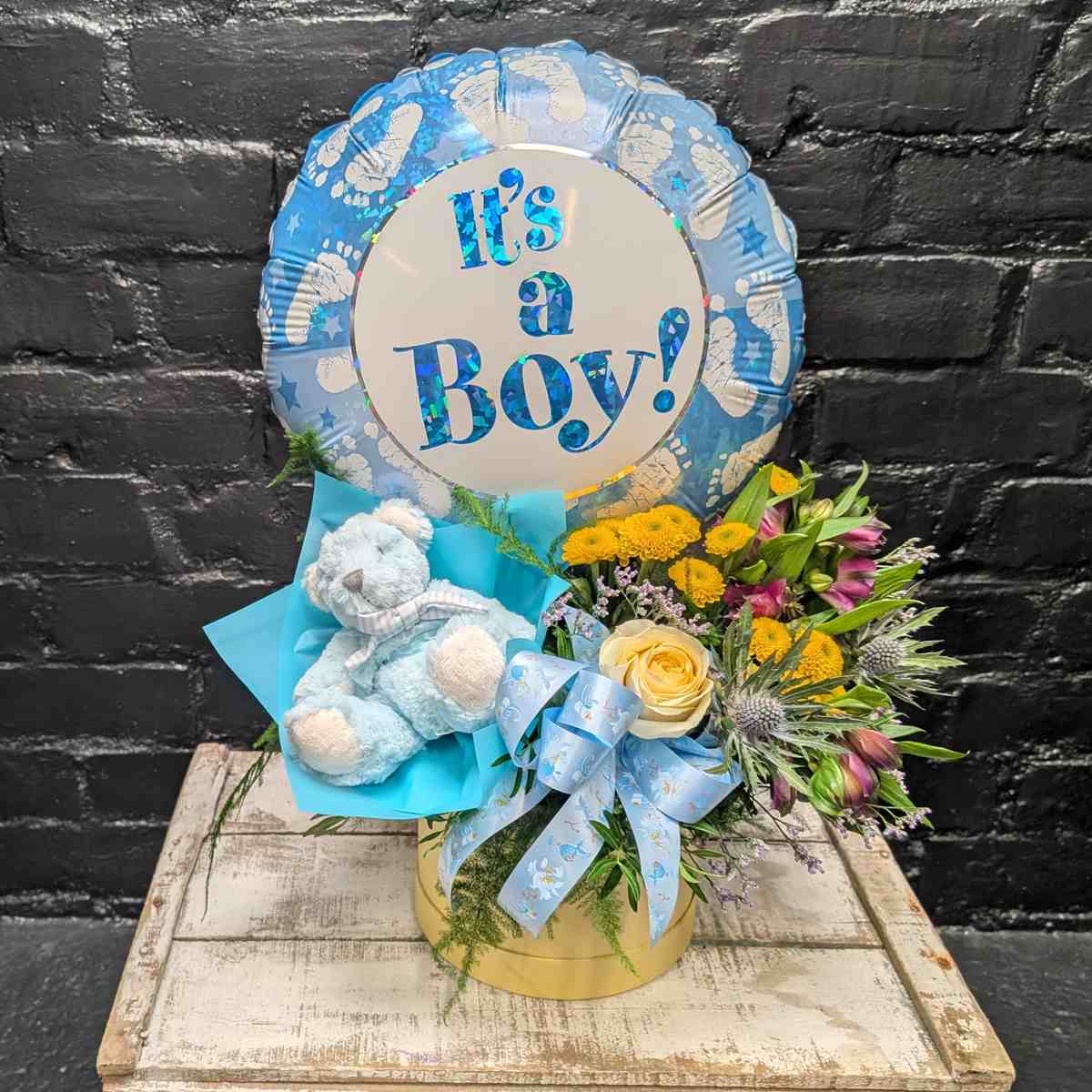 “It’s a Boy” new baby gift with blue balloon, soft blue teddy bear and yellow and purple flower arrangement in a gold hat box on a wooden crate – Anastasia Florists Aberdeen - Side View