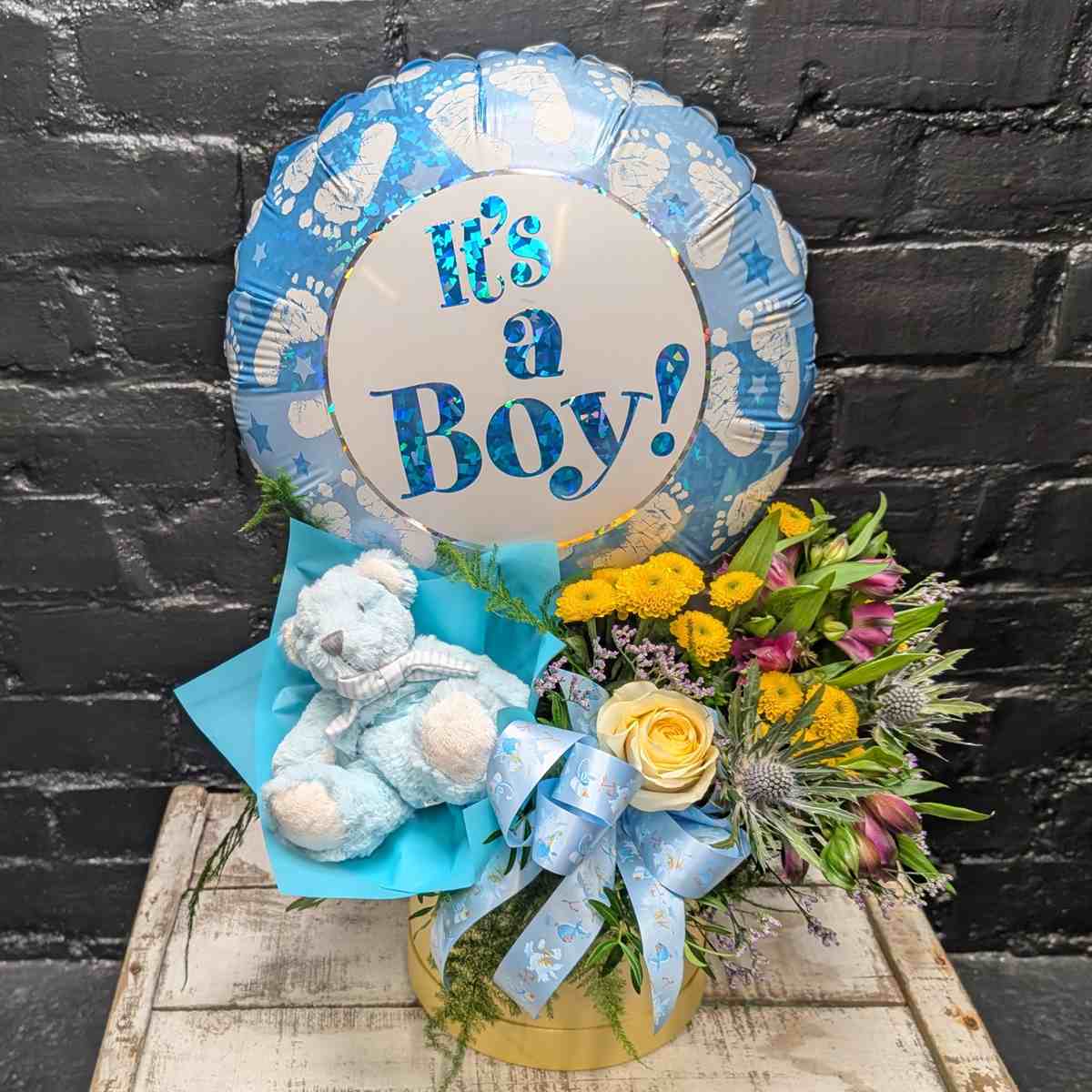 “It’s a Boy” new baby gift with blue balloon, soft blue teddy bear and yellow and purple flower arrangement in a gold hat box on a wooden crate – Anastasia Florists Aberdeen - Far View