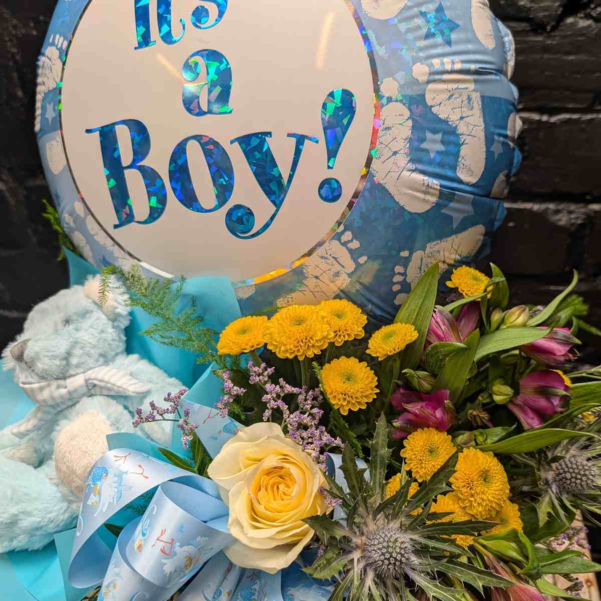 “It’s a Boy” new baby gift with blue balloon, soft blue teddy bear and yellow and purple flower arrangement in a gold hat box on a wooden crate – Anastasia Florists Aberdeen - close up