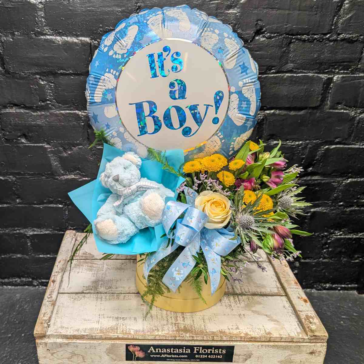 “It’s a Boy” new baby gift with blue balloon, soft blue teddy bear and yellow and purple flower arrangement in a gold hat box on a wooden crate – Anastasia Florists Aberdeen