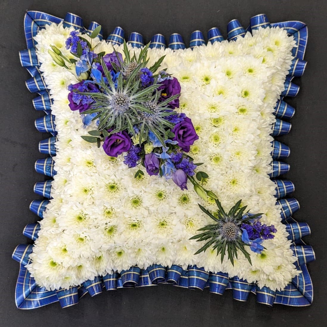 Blue tartan funeral cushion tribute with white chrysanthemum base and purple thistle spray