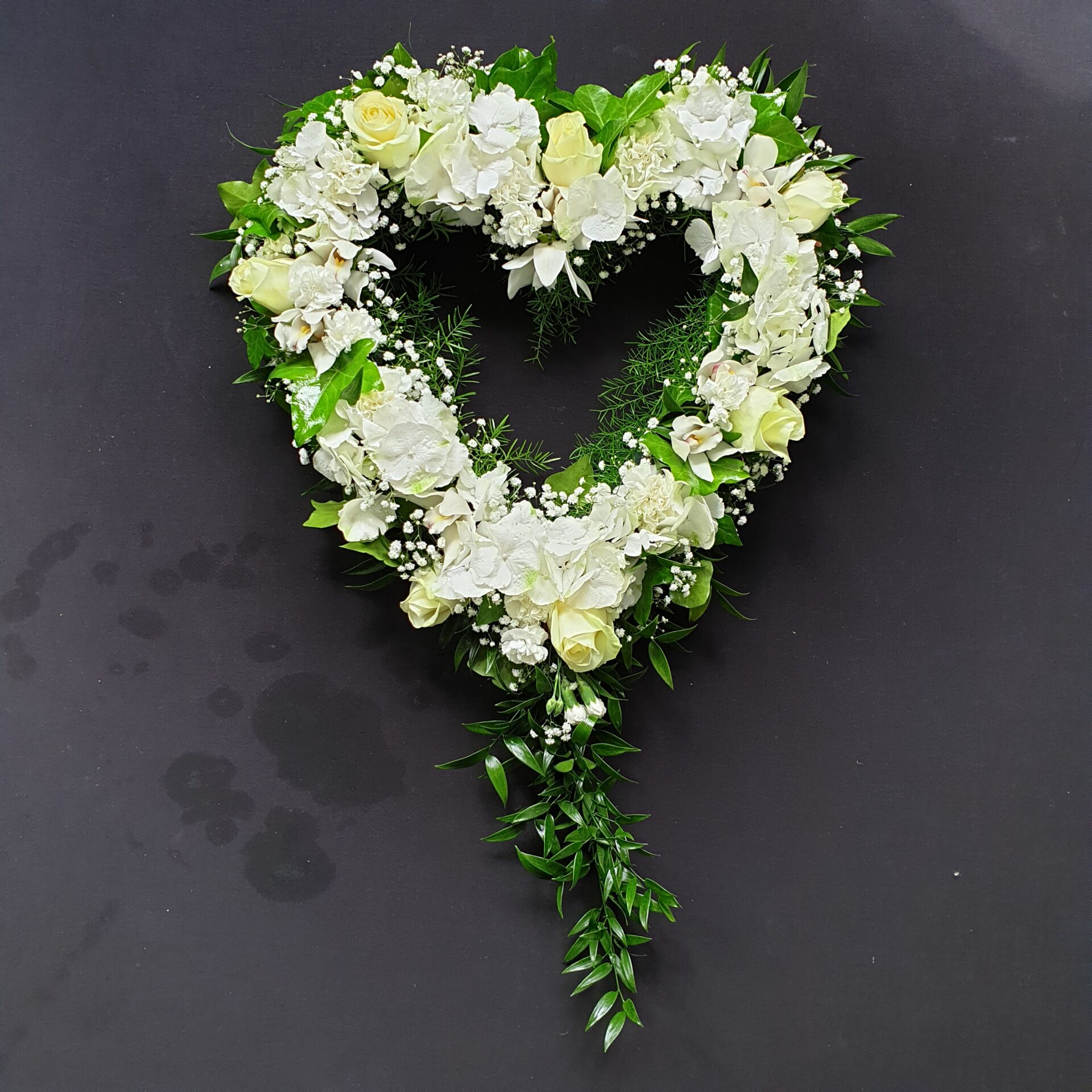White and green open heart funeral tribute with roses, hydrangea and trailing foliage on a dark background