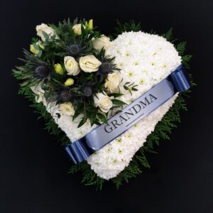 White chrysanthemum heart funeral tribute with ivory roses, blue thistle and blue Grandma ribbon on a black background