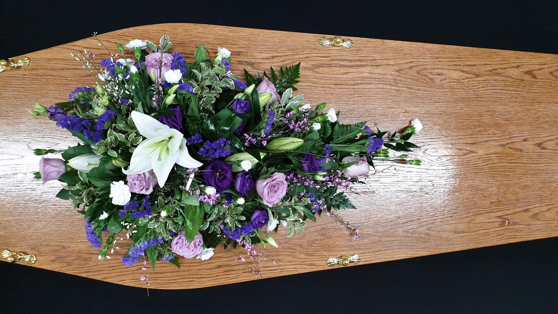 Lilac and purple lily single ended funeral spray on a wooden coffin with lisianthus and mixed greenery.