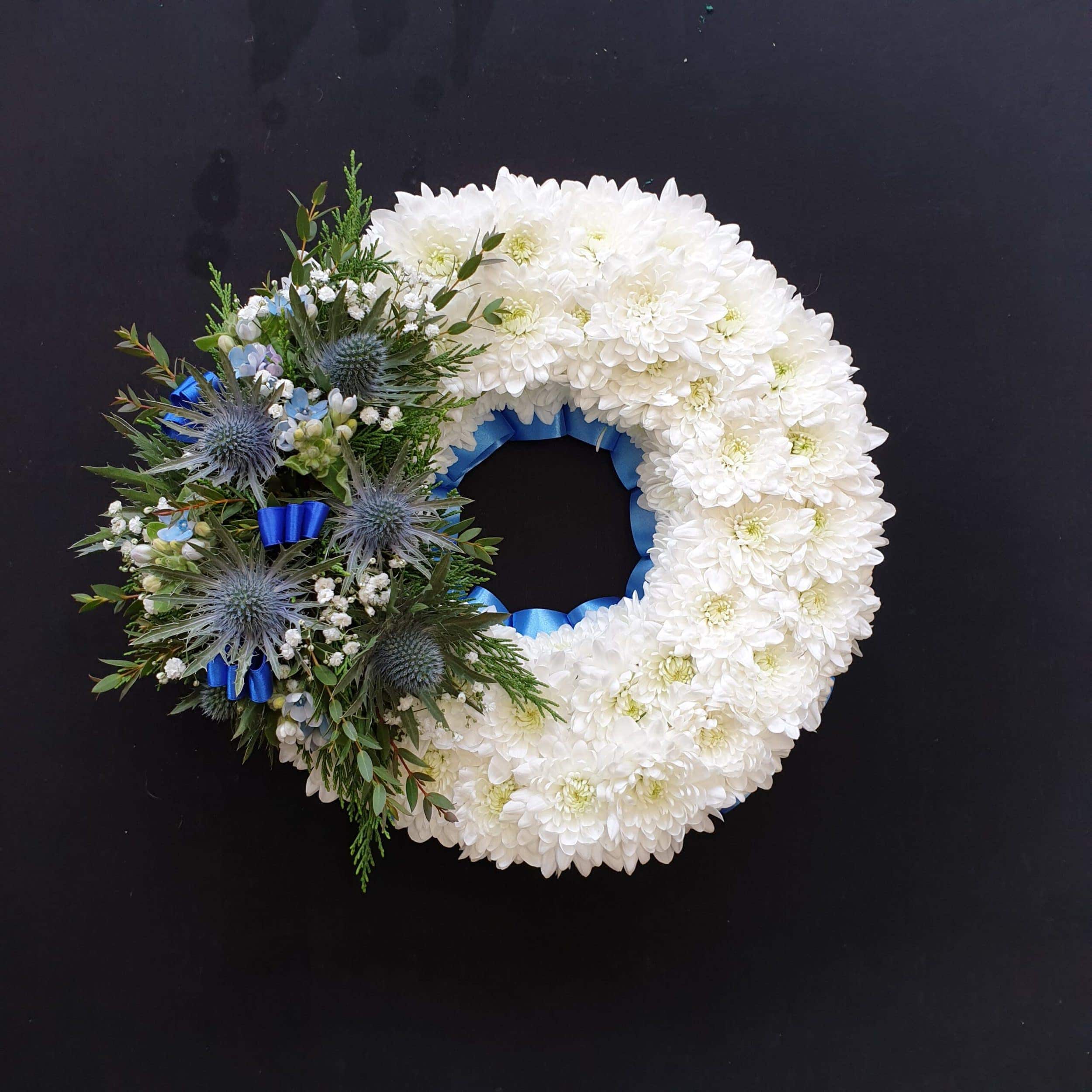 Miniature white chrysanthemum funeral wreath with blue thistles, gypsophila and foliage on a black background