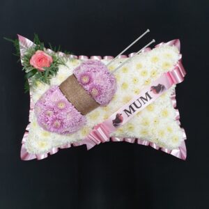 Mum knitting funeral pillow tribute with white chrysanthemum base, pink ribbon and wool with needles