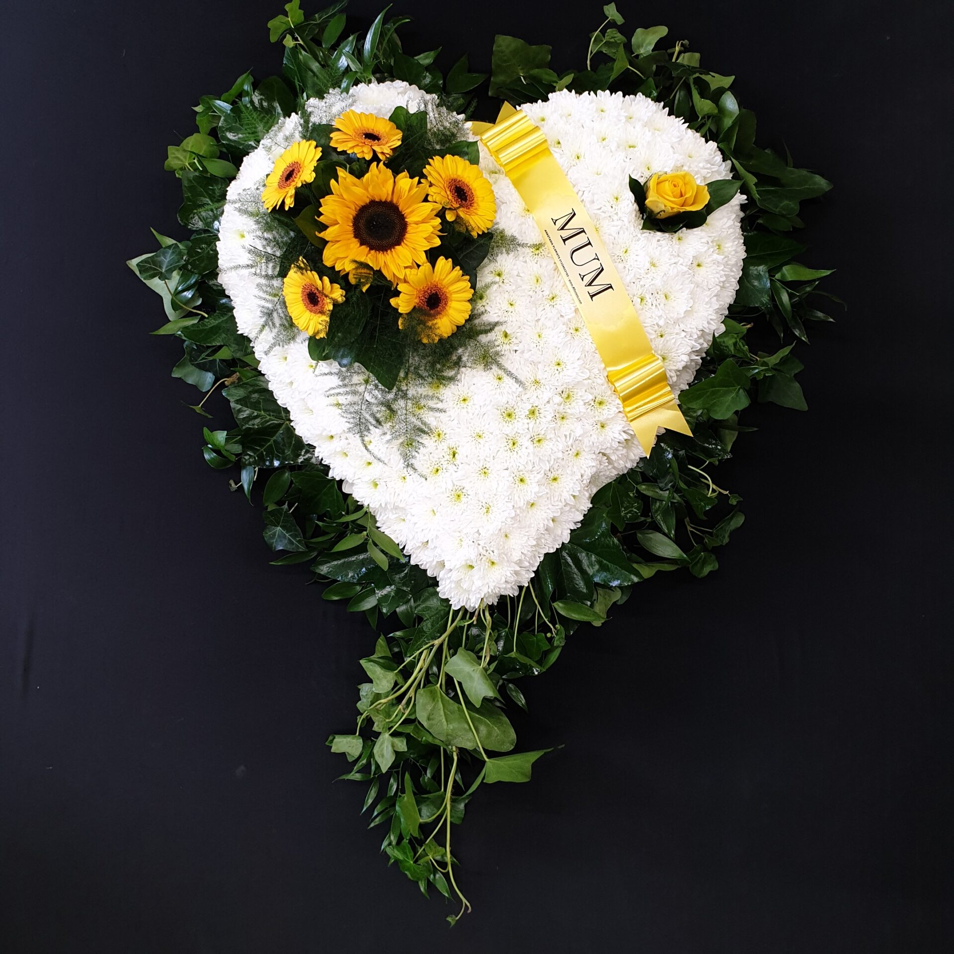 White chrysanthemum heart funeral tribute with yellow sunflowers, greenery and yellow Mum ribbon on a black background