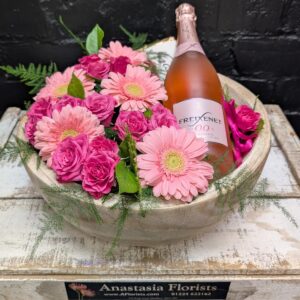 Pink flowers arranged in a wooden bowl with a bottle of sparkling drink.
