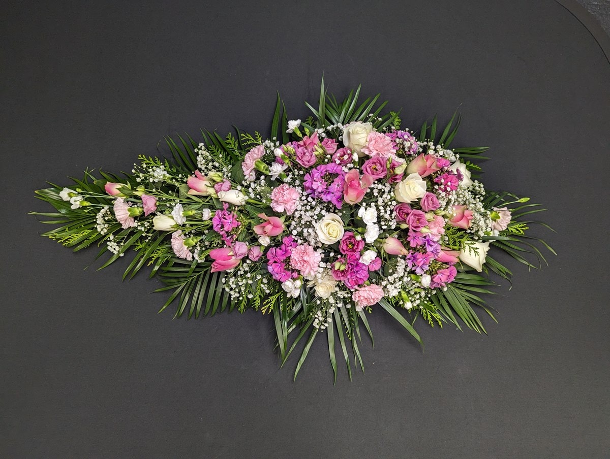 Pink and lilac single ended funeral spray with roses, carnations and gypsophila on a dark background.