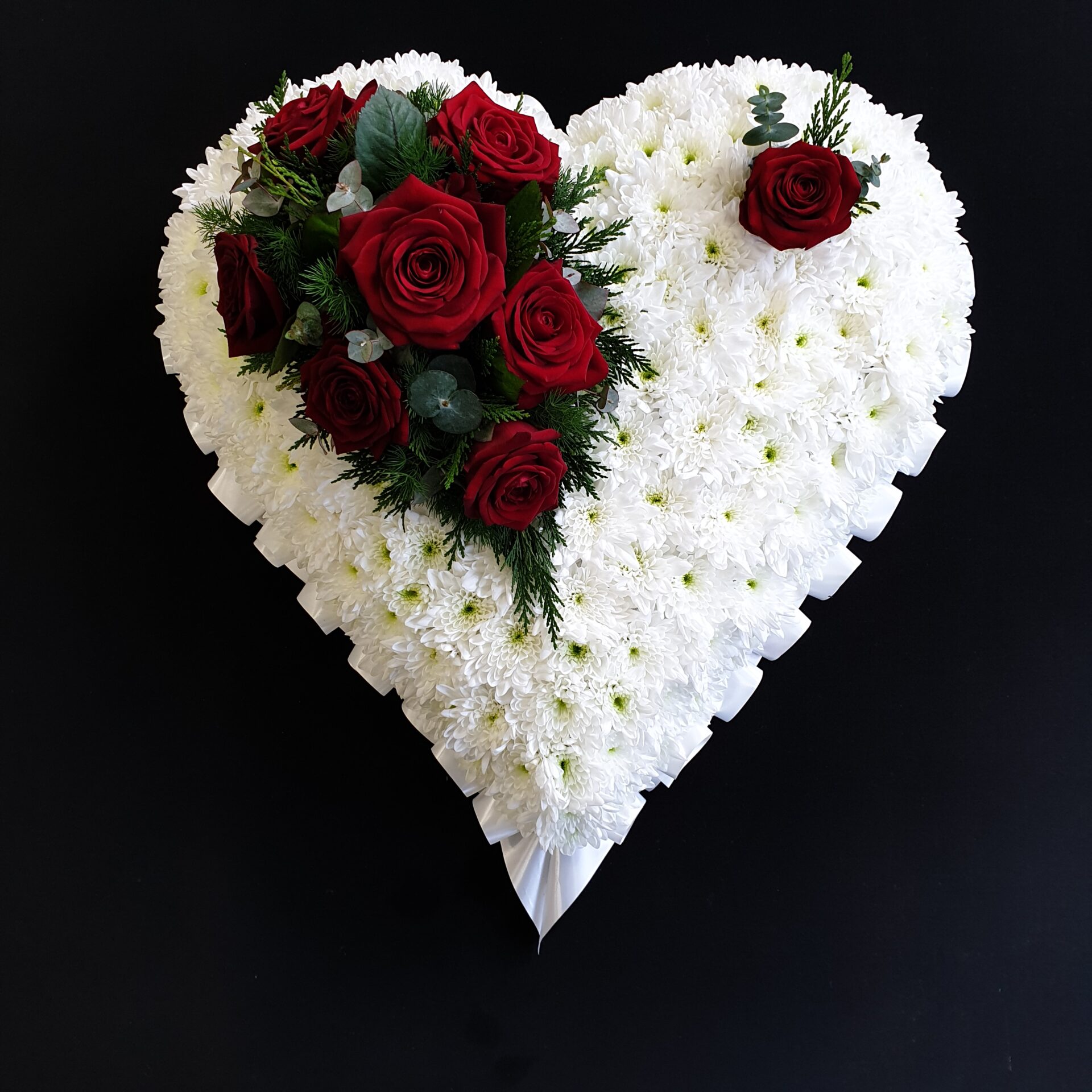 White chrysanthemum heart funeral tribute with red roses and greenery on a black background