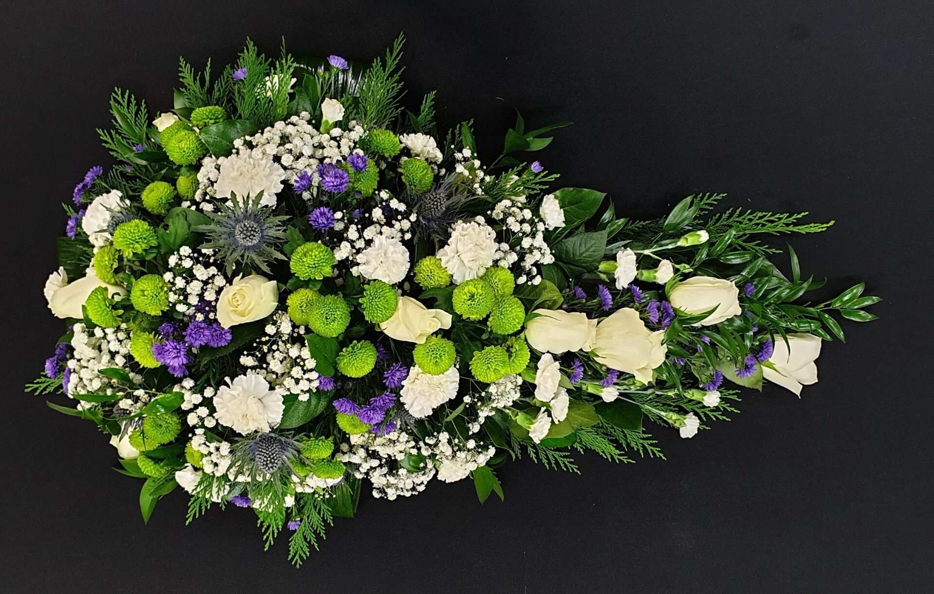 Thistle and lime green single ended funeral spray with ivory roses, white carnations and purple flowers on a black background.