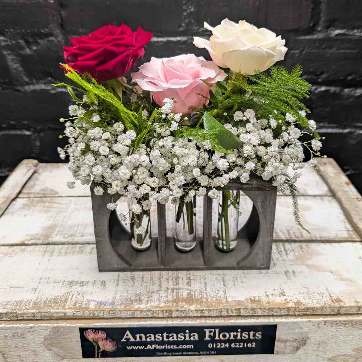 Three rose bud vase arrangement with red, pink and white roses, Gypsophila and greenery in a rustic wooden holder on a wooden crate – Anastasia Florists Aberdeen