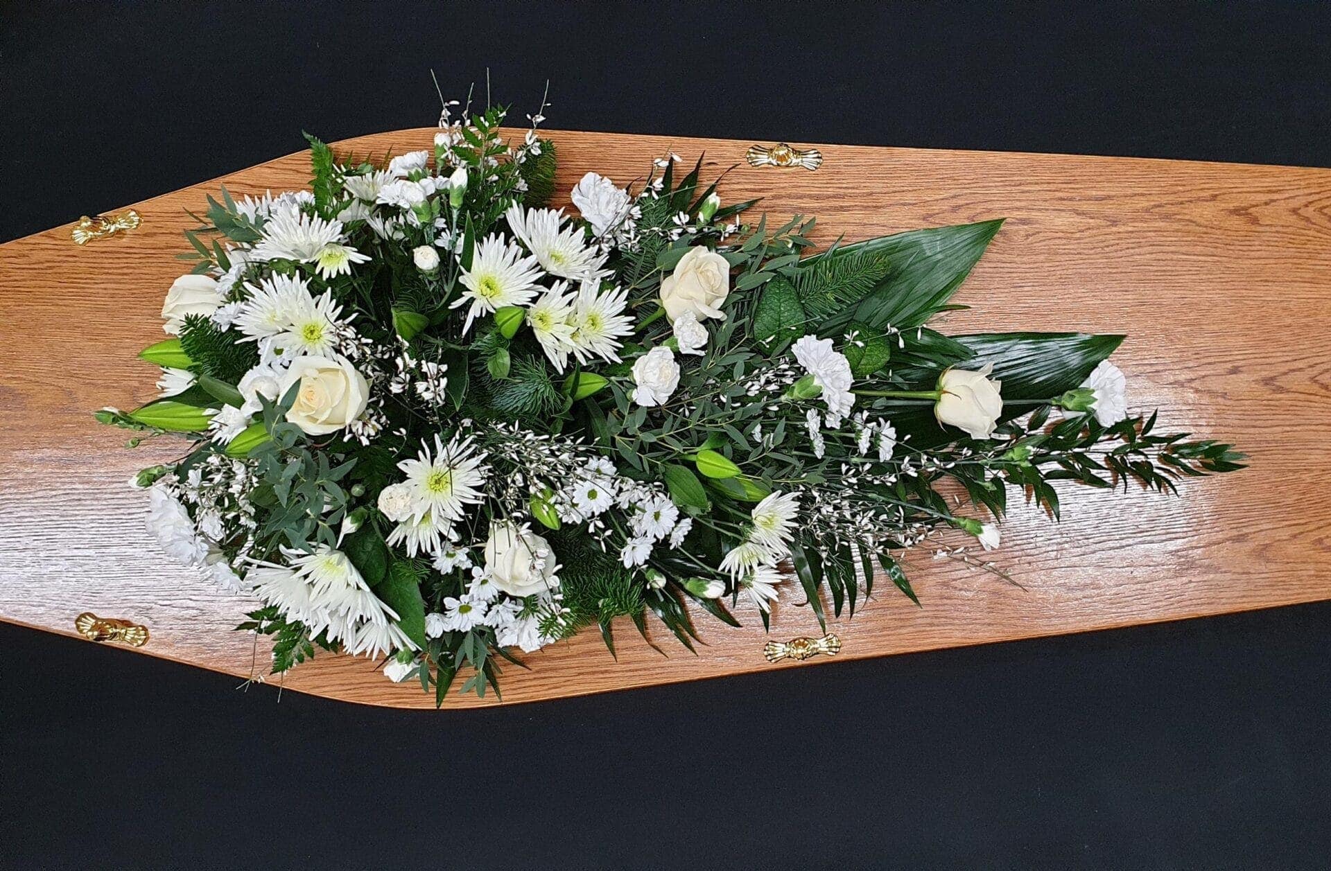White rose and chrysanthemum single ended funeral spray on a wooden coffin with carnations and mixed greenery.