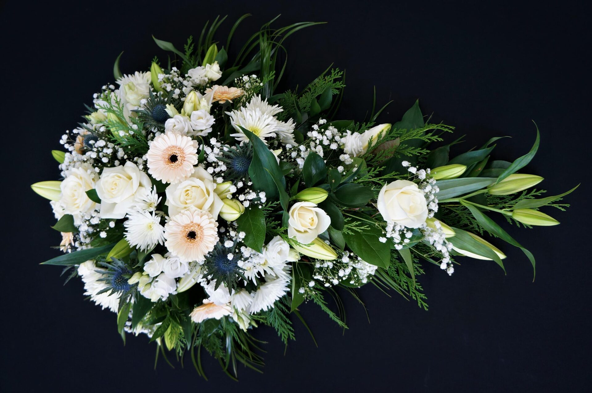 White rose and gerbera single ended funeral spray with lilies, gypsophila and greenery on a black background.