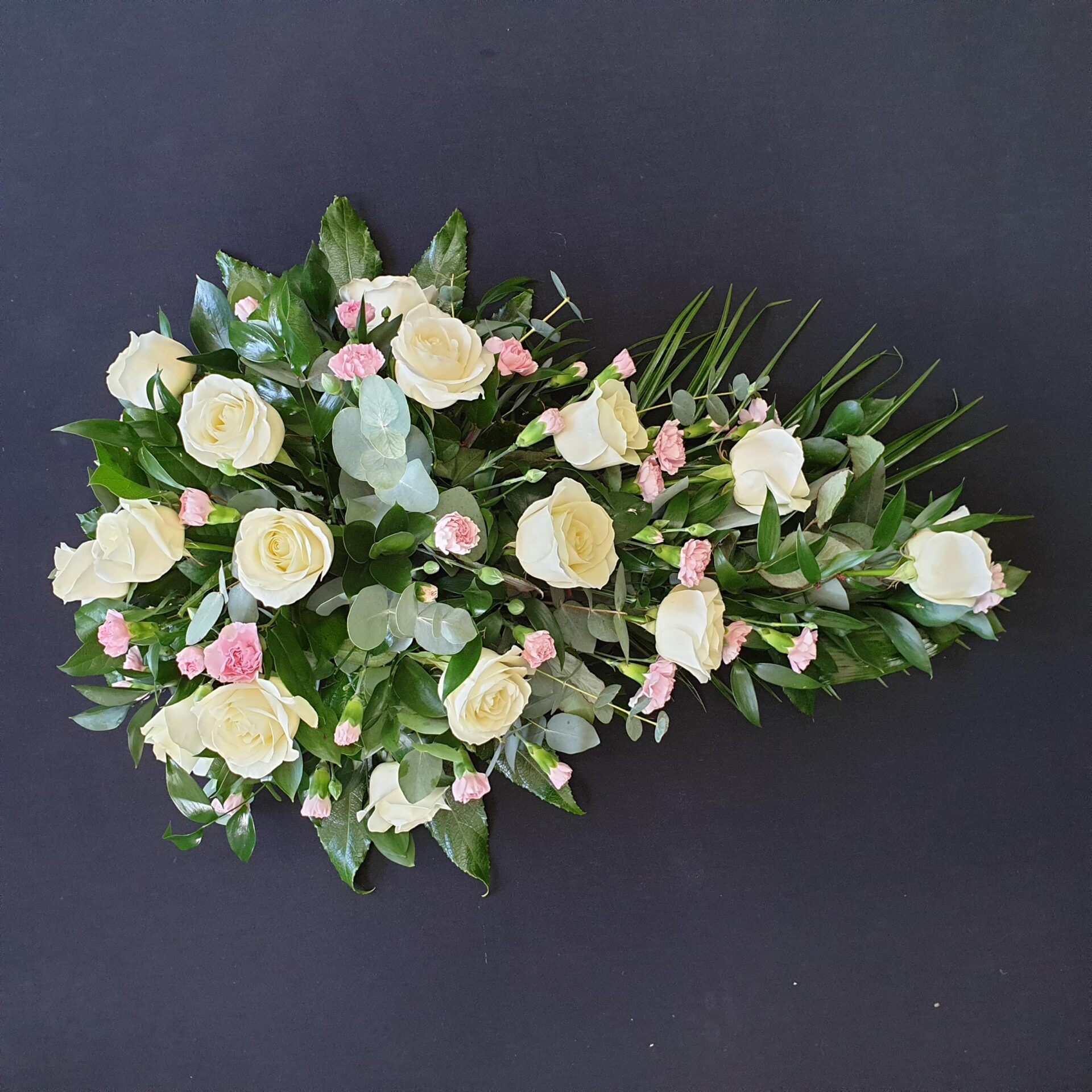 White rose and pale pink carnation single ended funeral spray with eucalyptus and greenery on a dark background.