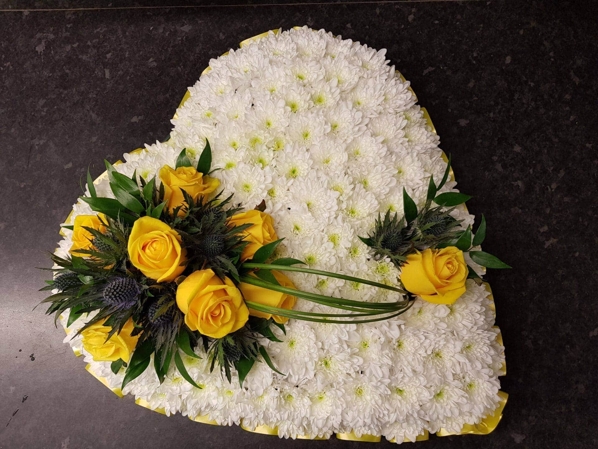 White chrysanthemum heart funeral tribute with yellow roses, thistle and yellow ribbon edging on a dark surface